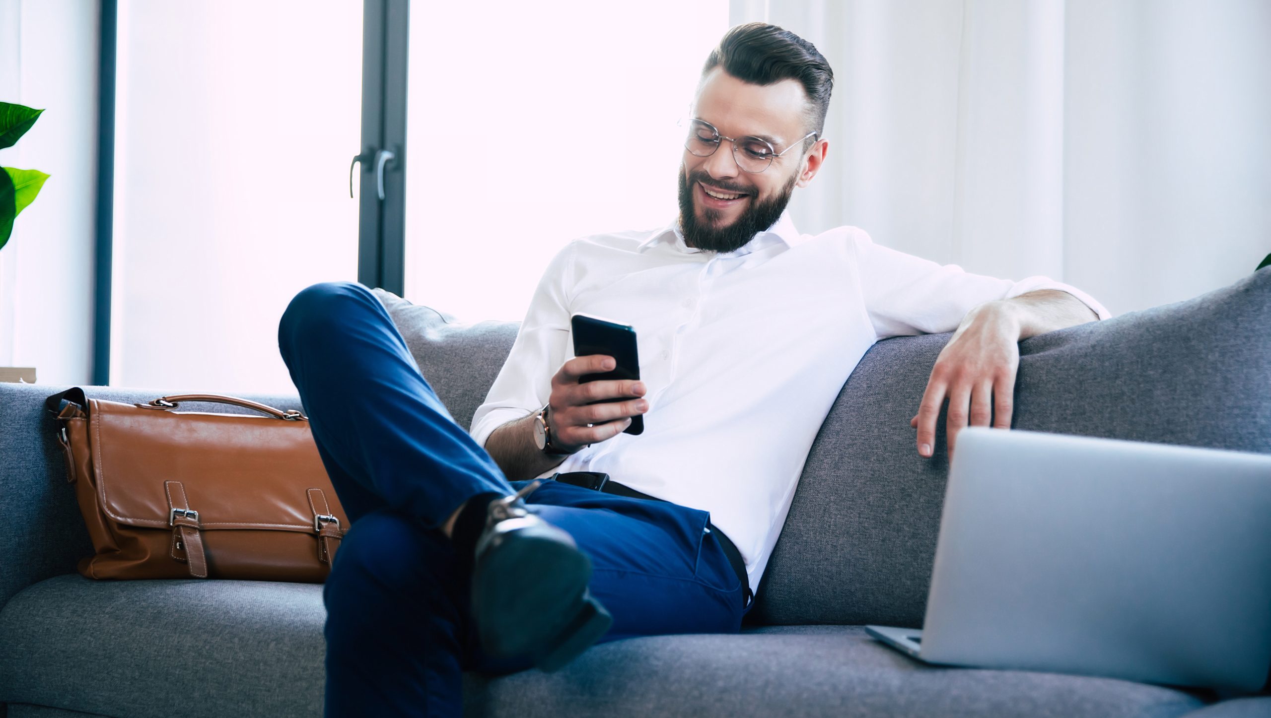 Ein junger Mann mit Brille sitzt entspannt neben seinem Laptop auf einem Sofa und betrachtet lächelnd den Screen seines Smartphones.